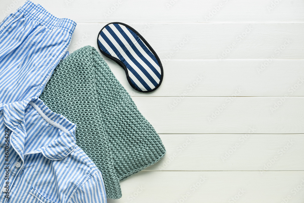 Sleep mask, pajamas and knitted blanket on white wooden background