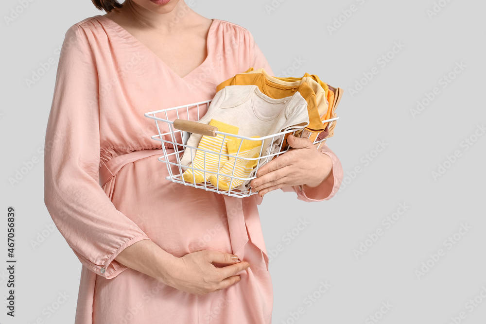 Young pregnant woman holding basket with baby clothes on light background