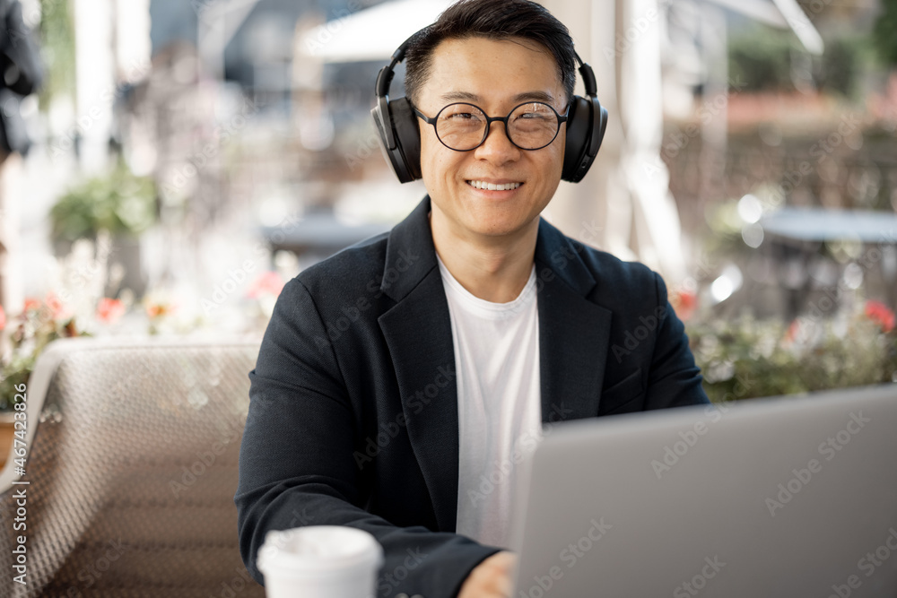 Asian businessman in earphones working on laptop in cafe. Concept of remote and freelance work. Smil