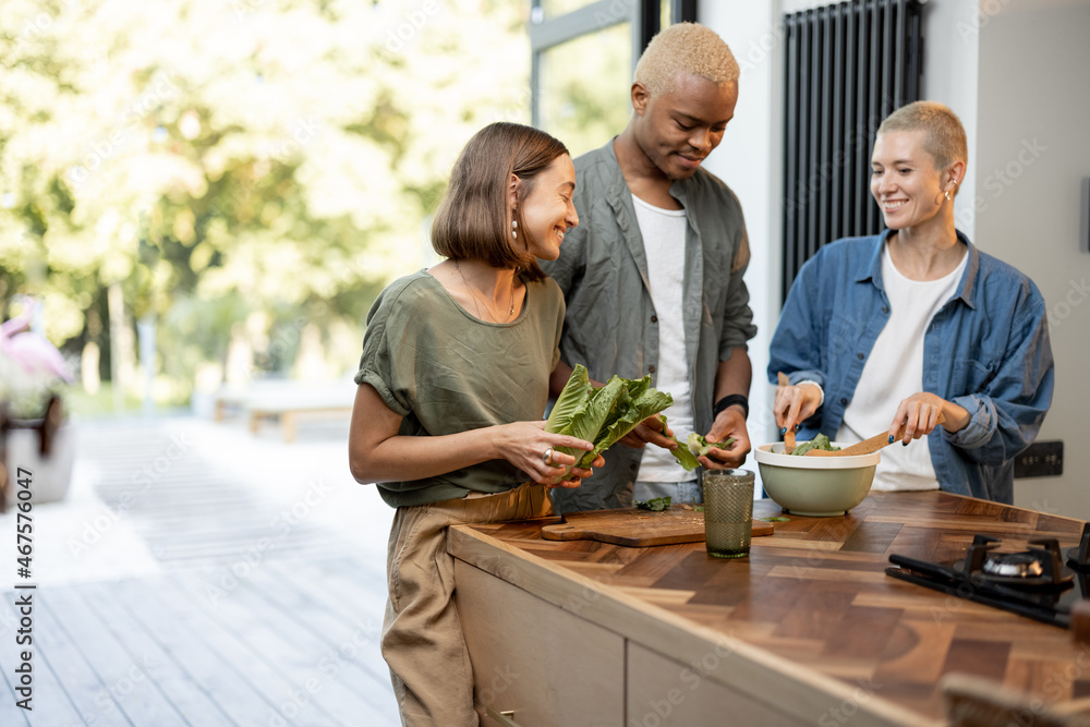 Friends cooking salad at home kitchen. Black man and european girls enjoying time together. Concept 