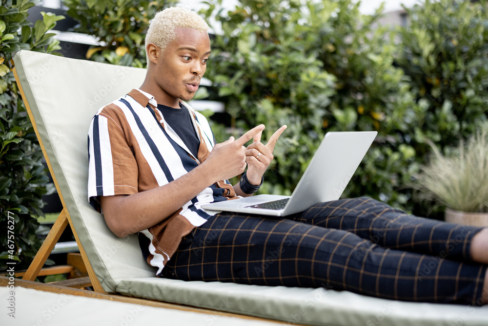 Latin man talking online or having zoom meeting using laptop computer while laying on lounger at hou