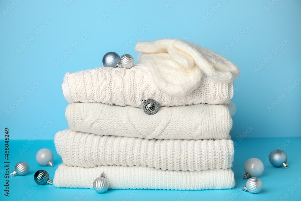 Stack of warm sweaters, mittens and Christmas balls on color background