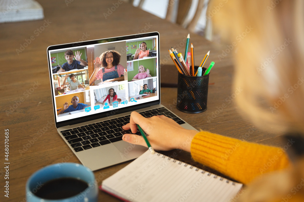 Caucasian woman using laptop for video call, with smiling diverse elementary school pupils on screen