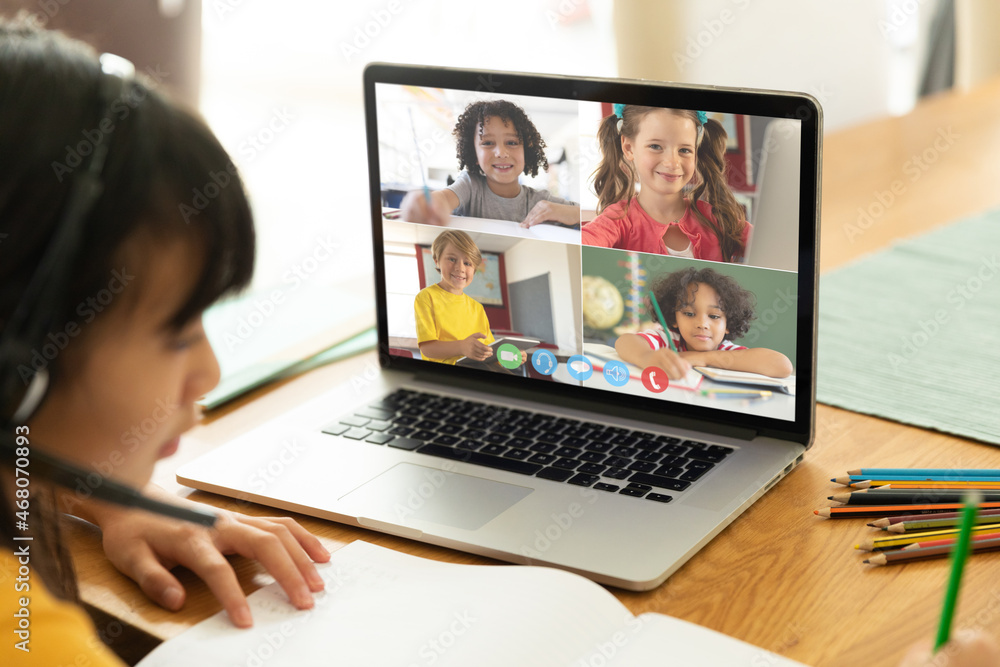 Asian girl using laptop for video call, with smiling diverse elementary school pupils on screen