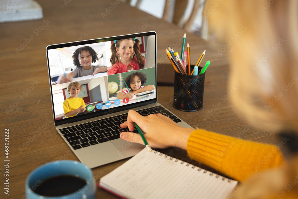 Caucasian woman using laptop for video call, with smiling diverse elementary school pupils on screen