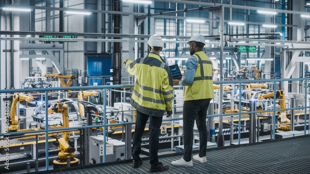 Two Car Factory Specialists in High Visibility Vests Using Laptop Computer. Engineers Discussing Aut