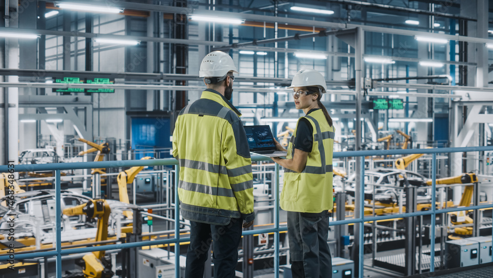 Male Specialist and Female Car Factory Engineer in High Visibility Vests Using Laptop Computer. Auto