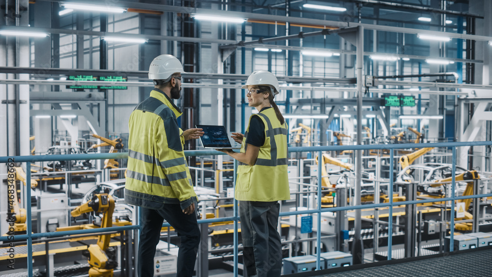 Male Specialist and Female Car Factory Engineer in High Visibility Vests Using Laptop Computer. Auto