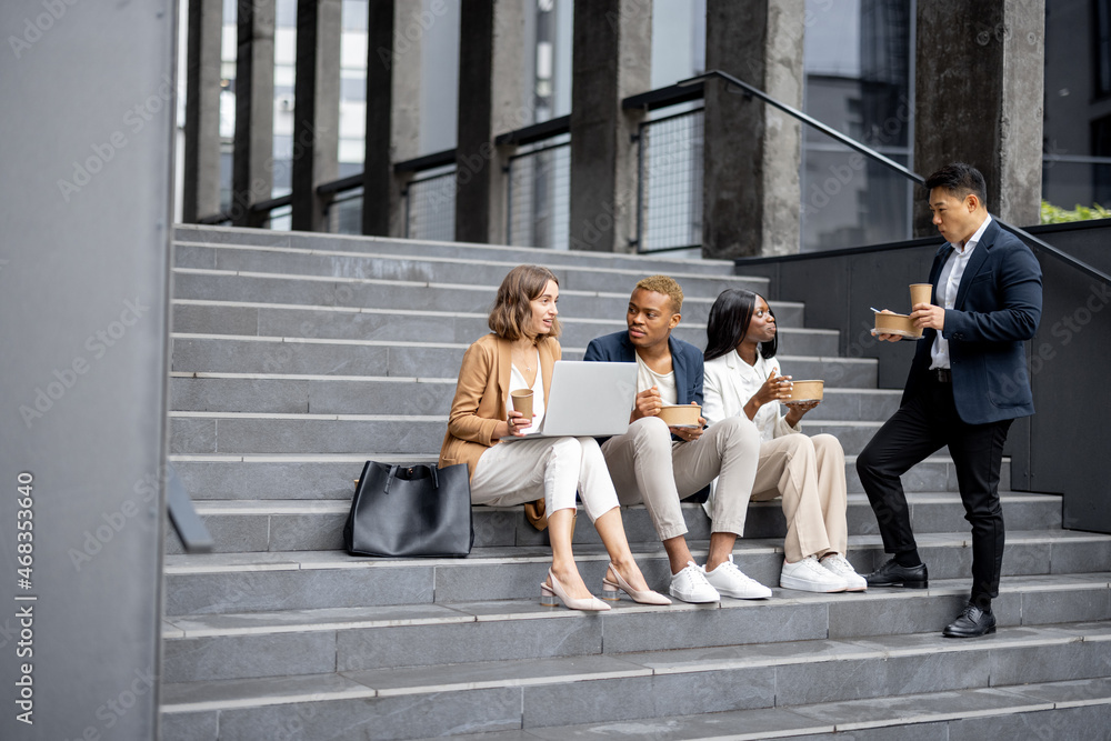 Multiracial businessteam talking, eating and drinking coffee while sitting on stairs in city. Concep