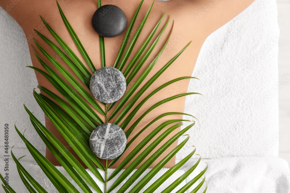 Young African-American woman with palm leaf and stones lying on couch in spa salon