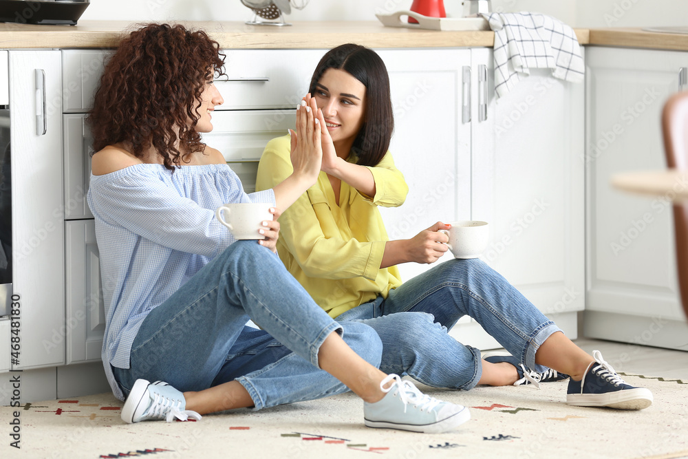 Young sisters sitting on floor, drinking tea and giving high five in kitchen