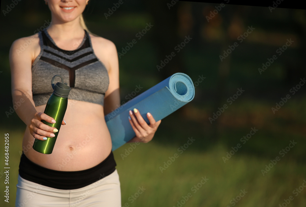 Young pregnant woman with yoga mat and bottle of water in park