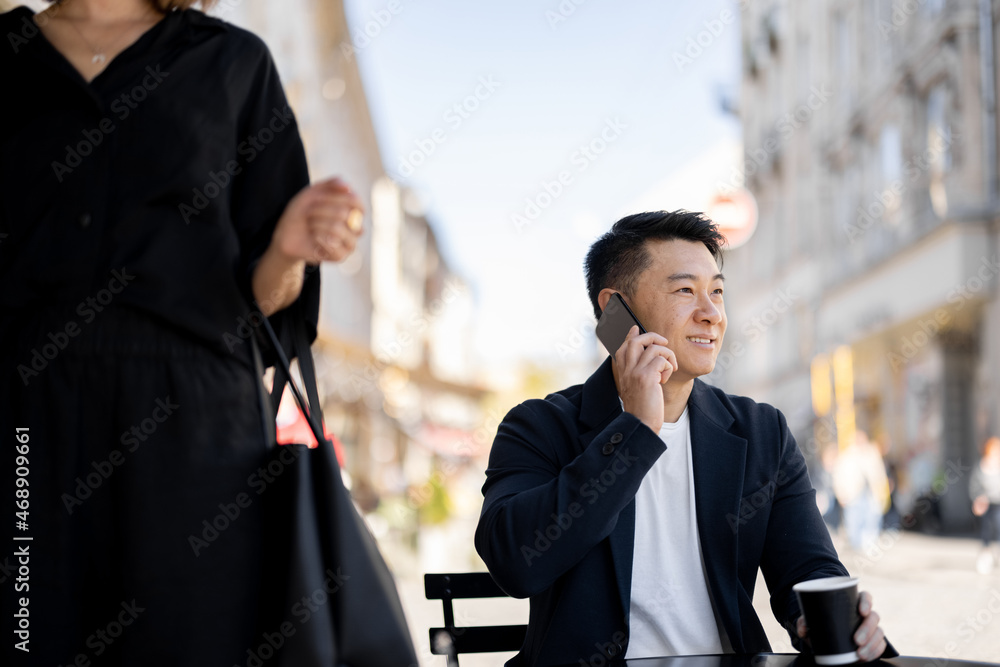 亚洲商人在户外咖啡馆用智能手机聊天。路过的女人。远程和频率的概念