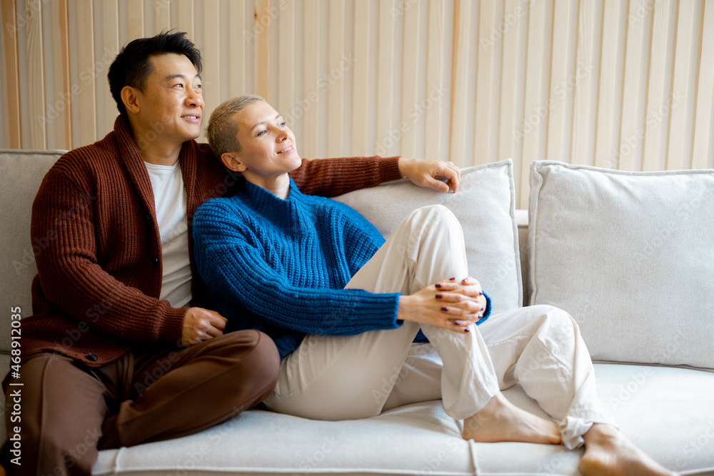 Asian man and young caucasian woman sitting and resting on sofa at home. Concept of relationship and