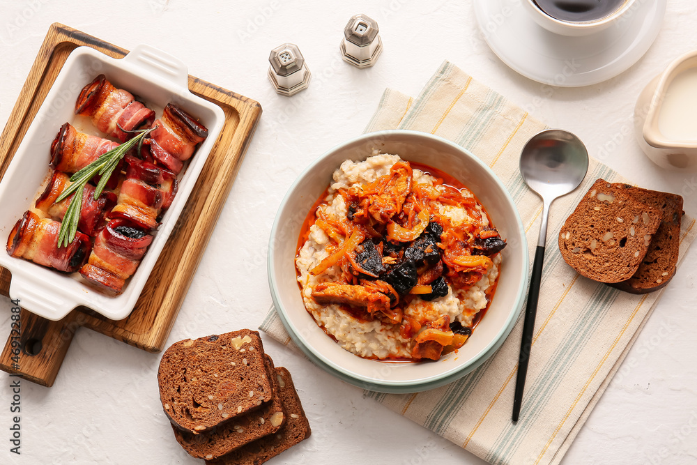 Bowl with delicious oatmeal and baking dish with tasty prunes baked in crispy bacon on light table