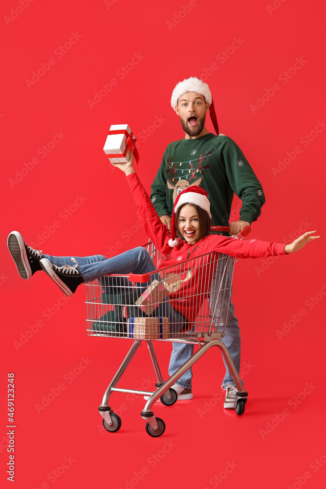 Happy young couple with shopping cart and Christmas gifts on color background