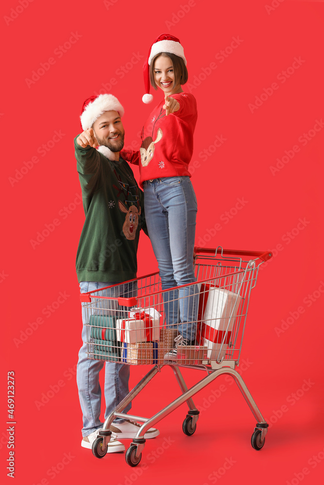 Happy young couple with shopping cart and Christmas gifts pointing at viewer on color background