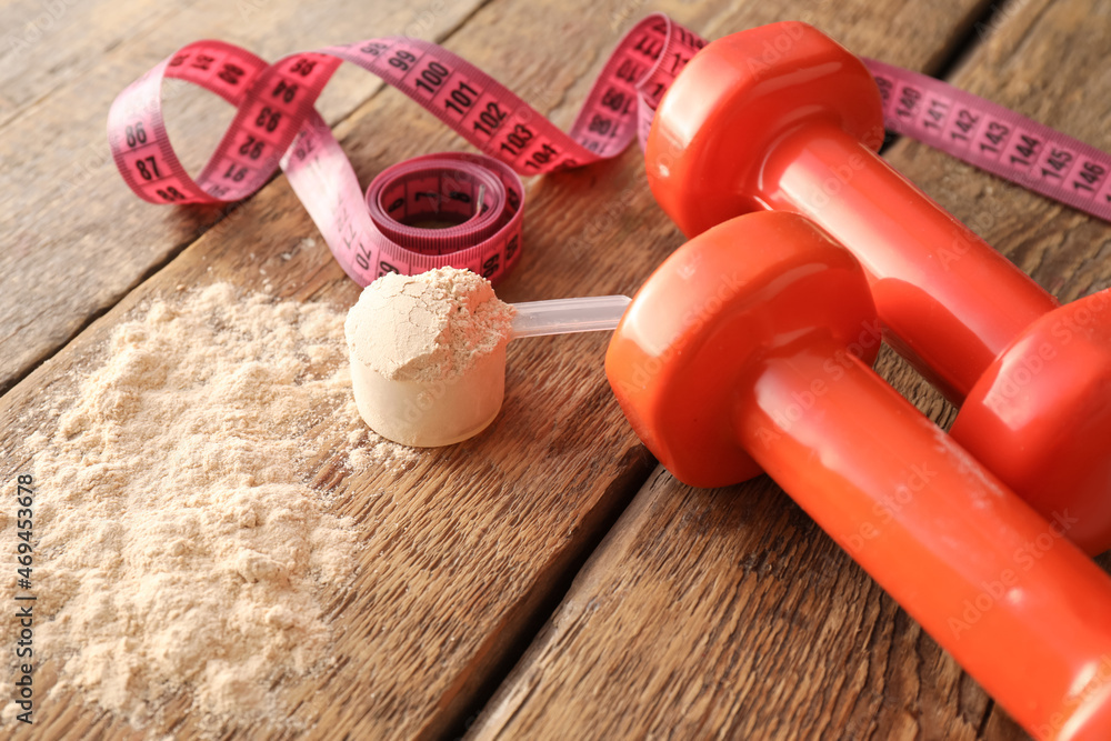 Scoop with protein powder, measuring tape and dumbbells on wooden table, closeup