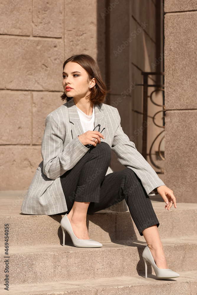 Fashionable woman with stylish accessories sitting on stairs and holding eyeglasses outdoors