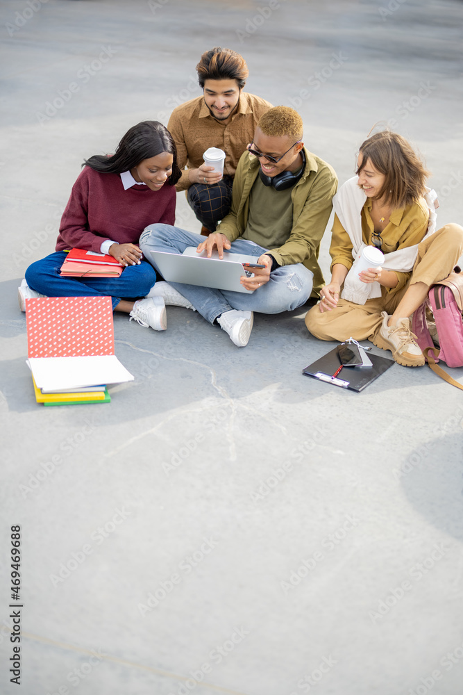 Multiracial students sitting and watching something on laptop computer on asphalt at university camp