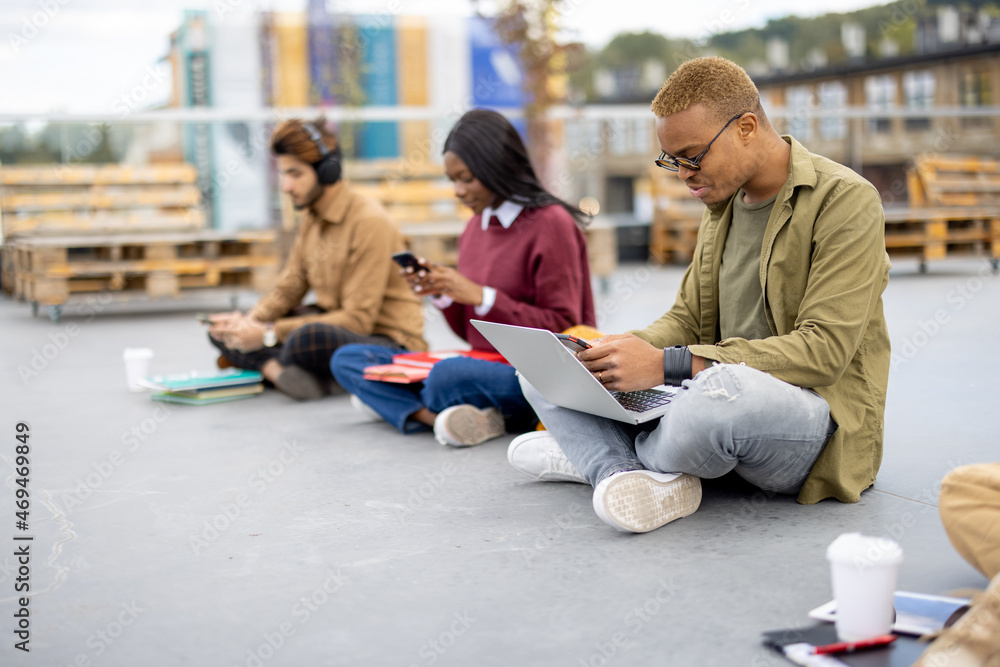 Row of multiracial students sitting and using smartphones on asphalt at university campus. Concept o