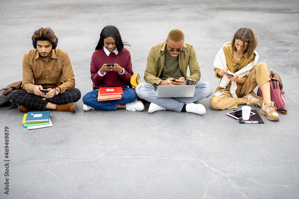 Multiracial students with digital devices sitting on asphalt and looking at camera at university cam