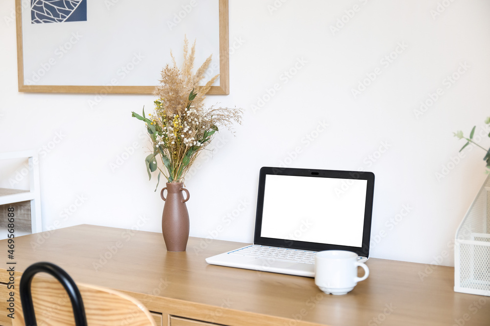 Table with laptop, cup and dried flowers in vase near light wall