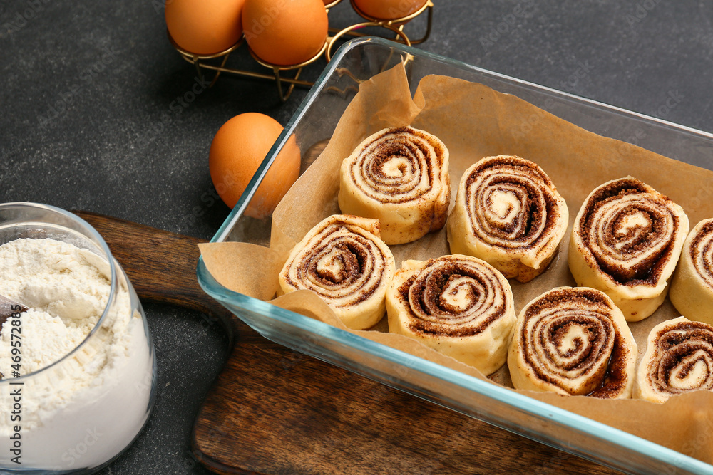 Baking dish of uncooked cinnamon rolls and ingredients on black background, closeup