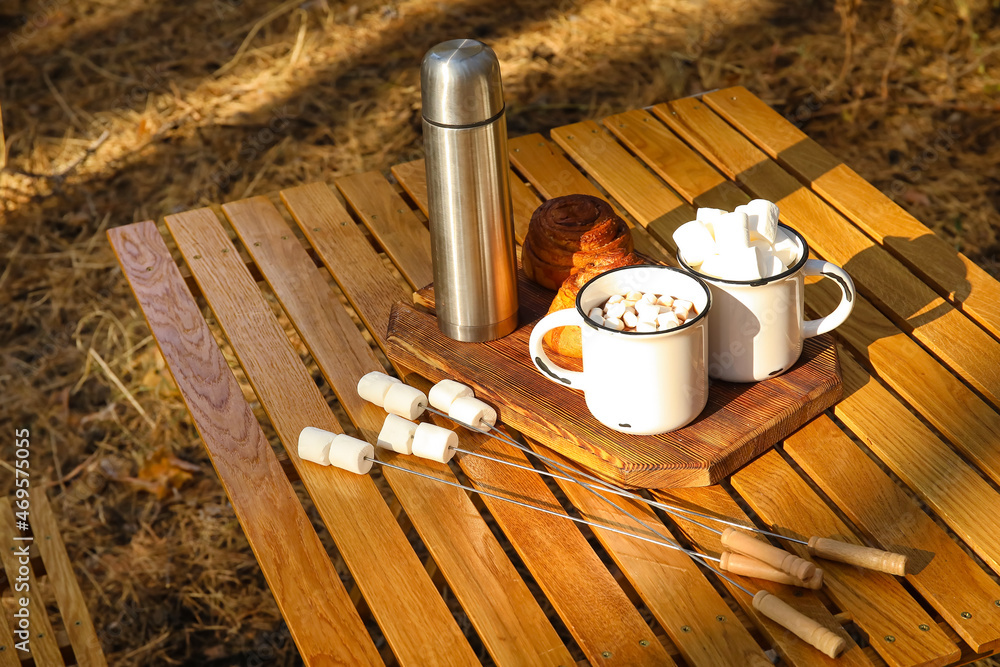 Cups of hot chocolate with tasty marshmallows and buns on table in autumn forest