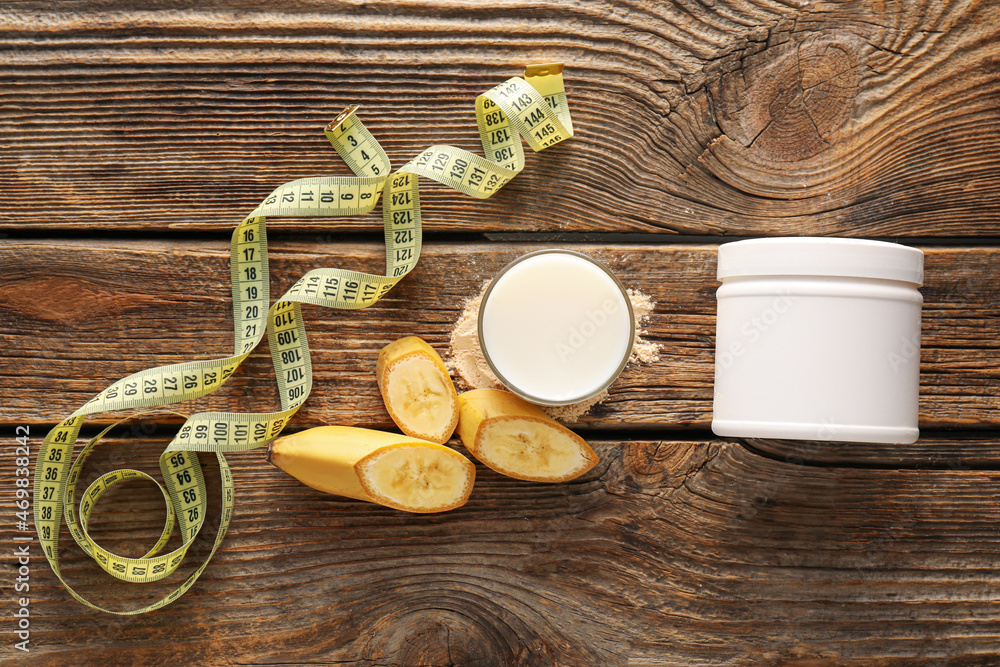 Glass of protein shake, jar with powder, sliced banana and measuring tape on wooden background