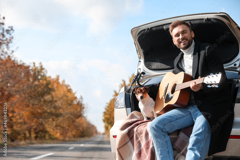 Handsome man with cute dog playing guitar in car trunk on autumn day