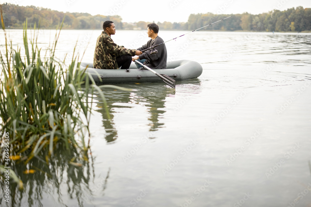 Multiracial male friends fishing with fishing rods on rubber boat in lake or river. Concept of rest,