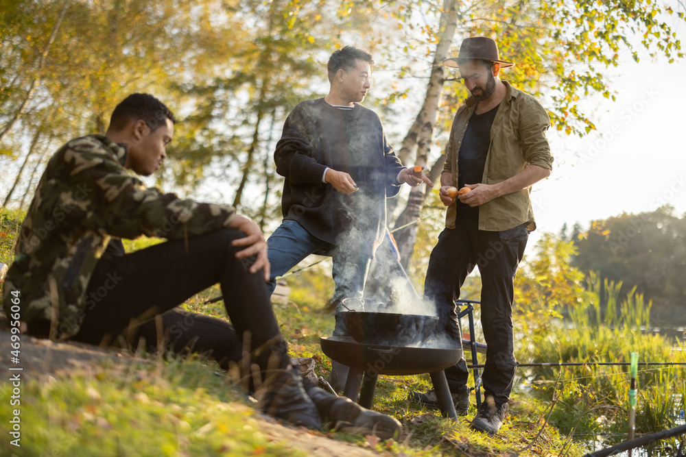 Multiracial male friends fishing and cooking on river or lake coast. Concept of rest, weekend and va