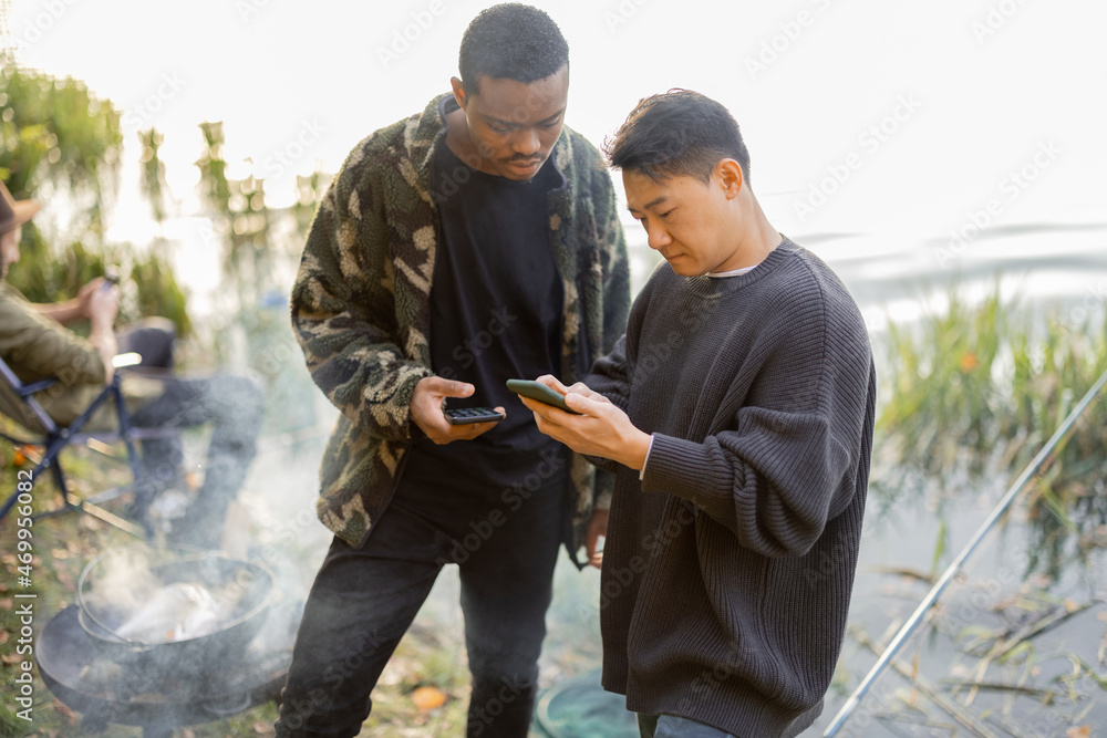 Asian and latin men using smartphones during resting on river or pond coast. Men fishing and cooking