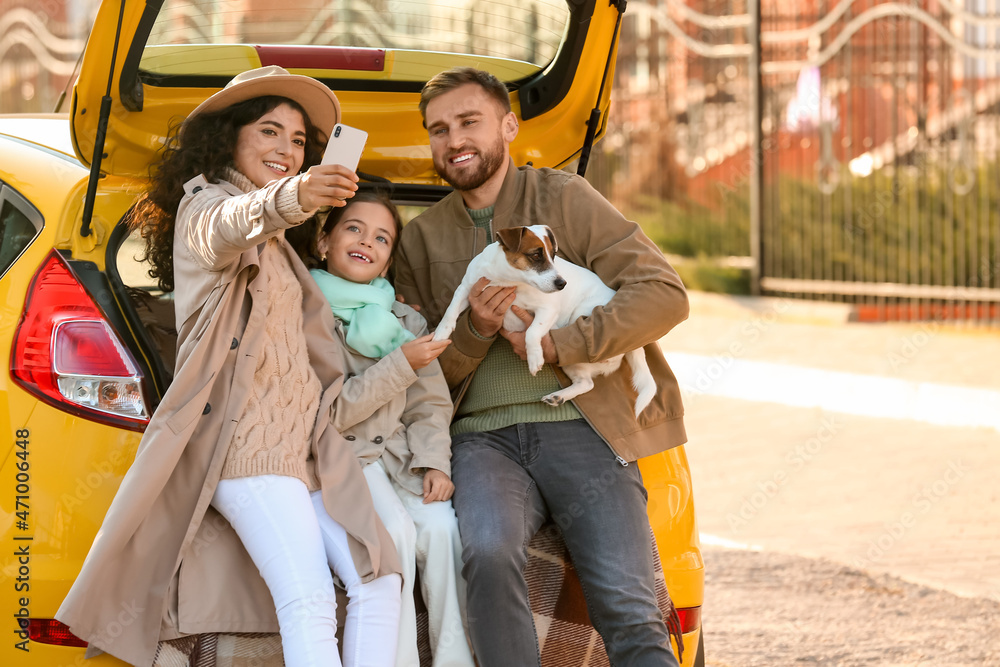 Happy parents with little daughter and cute dog taking selfie in car trunk outdoors