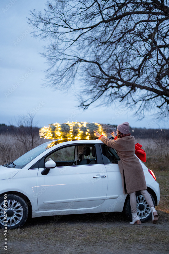 Woman putting illuminated Christmas tree on a rooftop of her small white car outdoors. Concept of pr