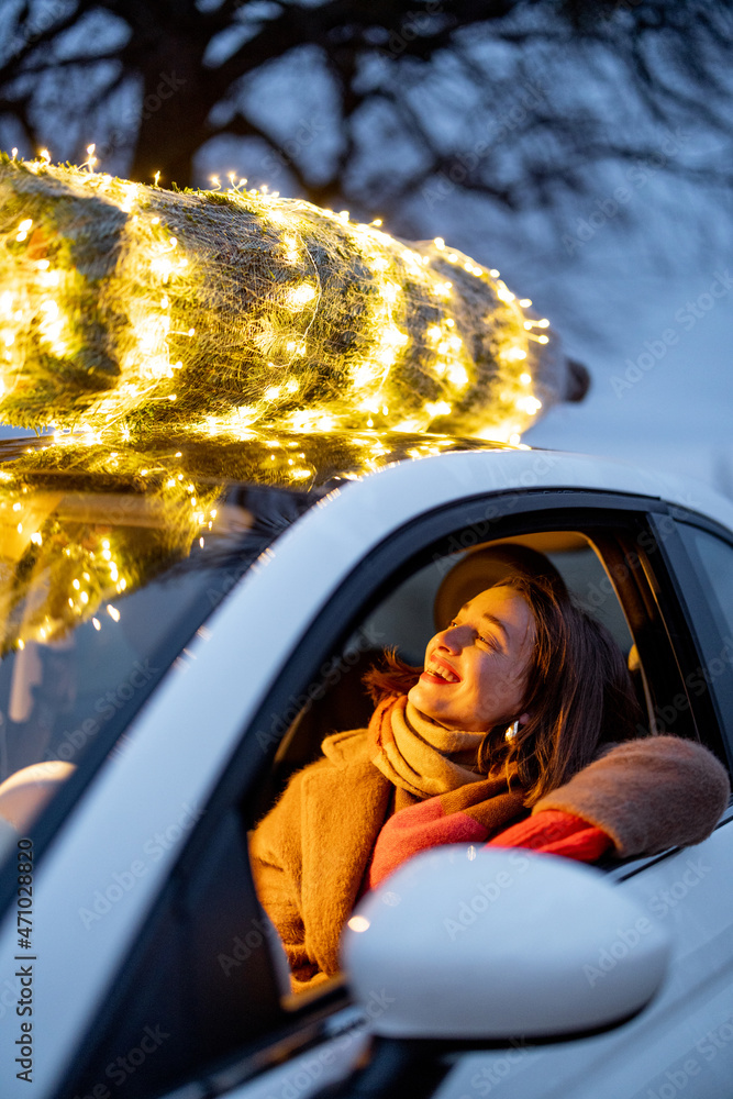 Happy woman driving car with a illuminated Christmas tree on a rooftop. Concept of New Year preparat