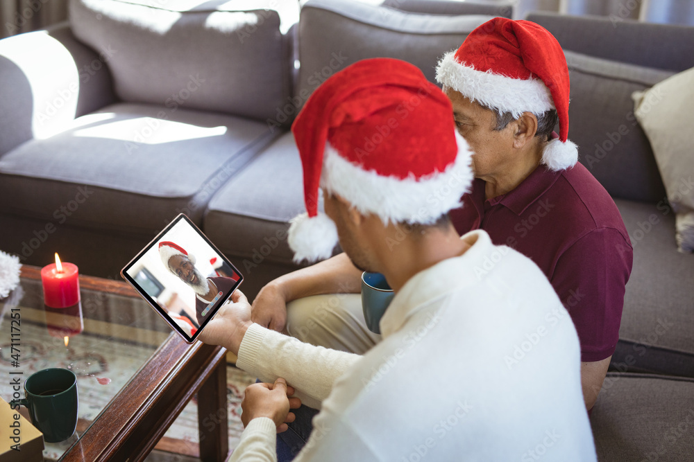 Biracial father and adult son in santa hats making tablet christmas video call with senior man