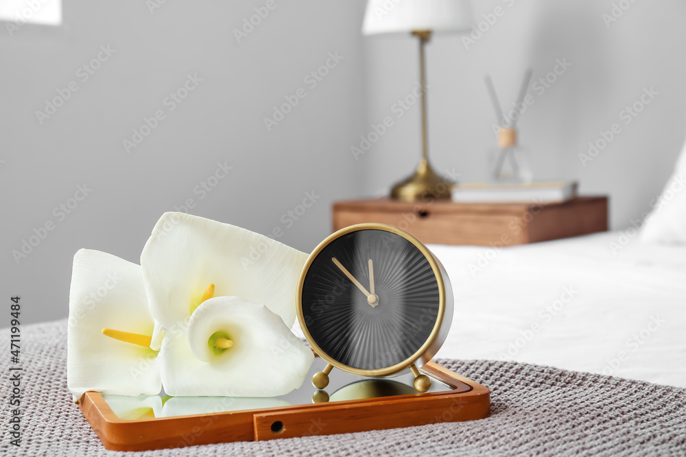 Stylish alarm clock with flowers on bed in room