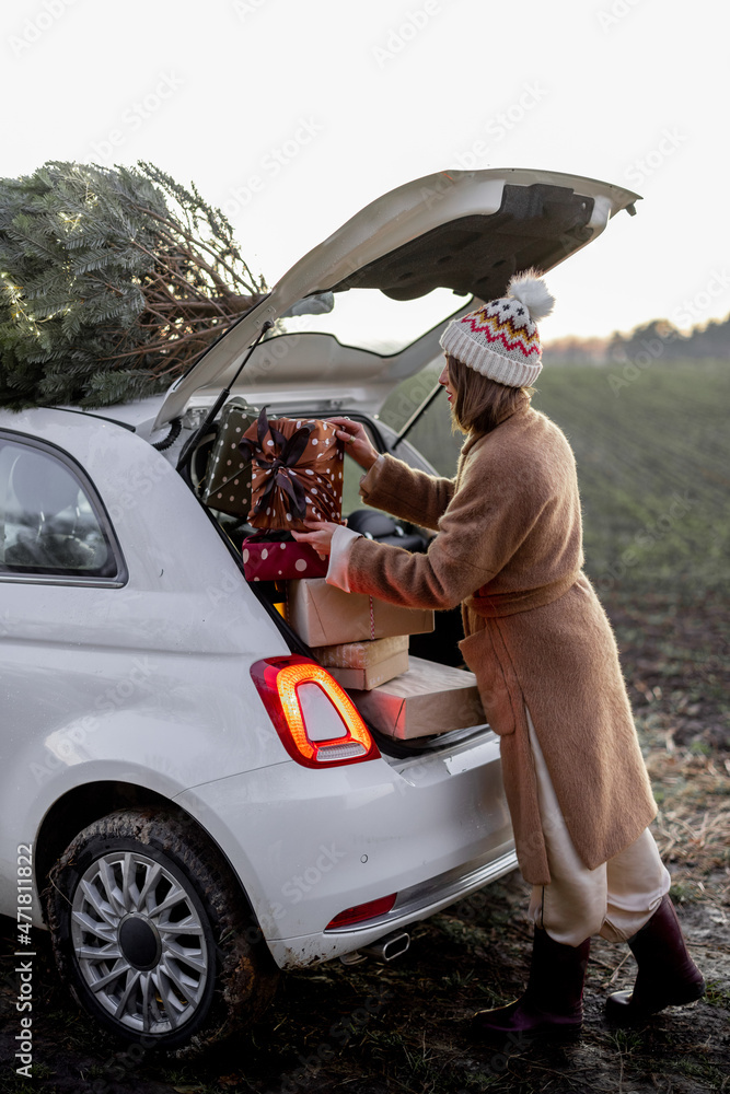 Woman packing gifts into the car with Christmas tree on a rooftop on nature at dusk. Getting ready f