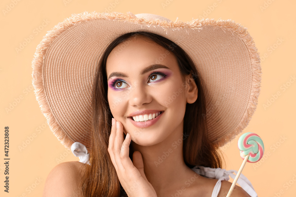 Beautiful young woman in hat with sweet lollipop on beige background