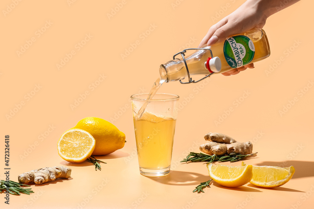 Woman pouring from bottle fresh kombucha with lemon and ginger into glass on color background