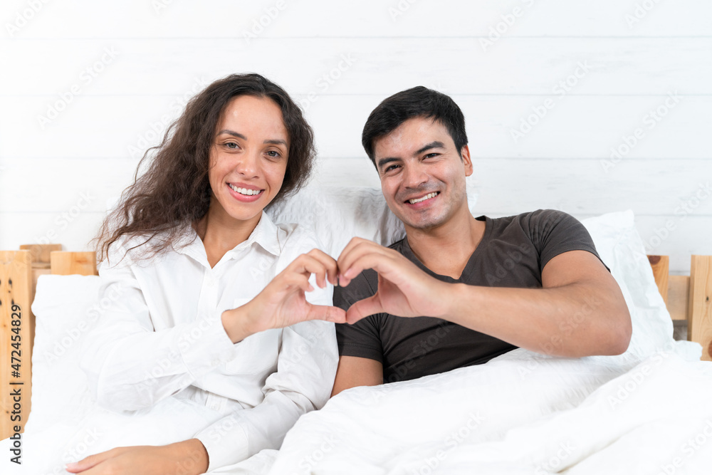 Happy Hispanic young couple making a heart shape with hands looking at the camera on the bed