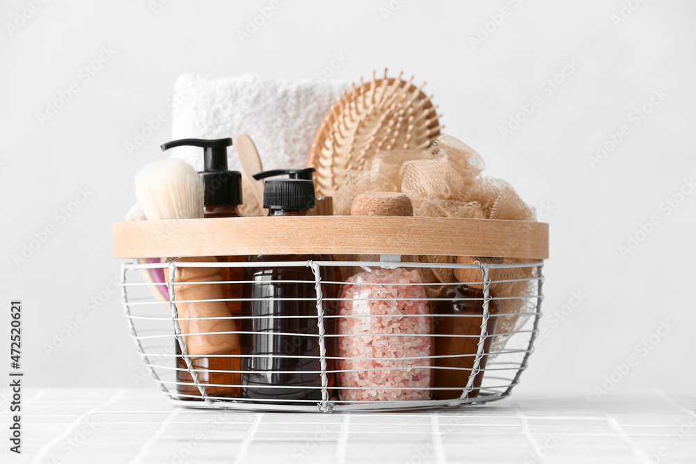 Basket with towel, bath supplies, hair brush and sponge on tile table against light background