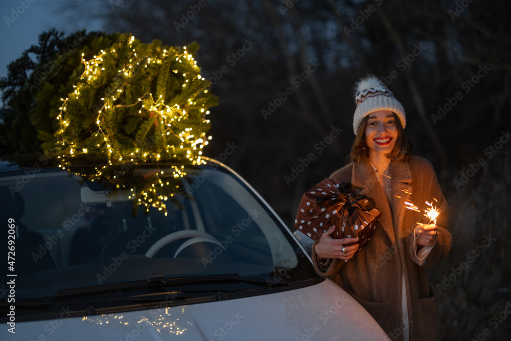 Happy caucasian woman firing sparklers near car with Christmas tree on nature at dusk. Concept of ce