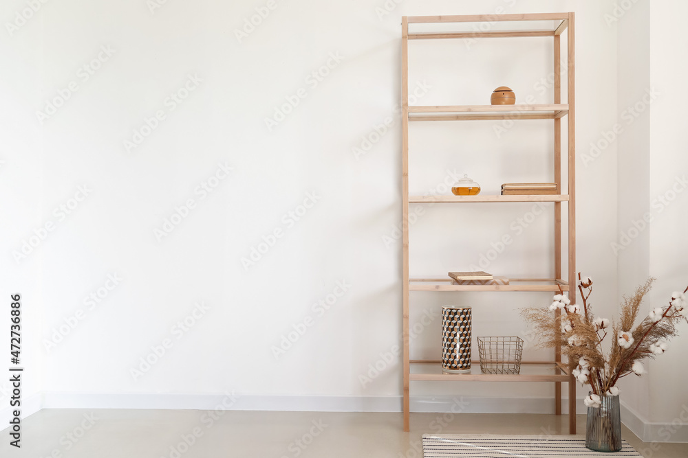 Bookcase with teapot, pampas grass and cotton branches in vase near light wall