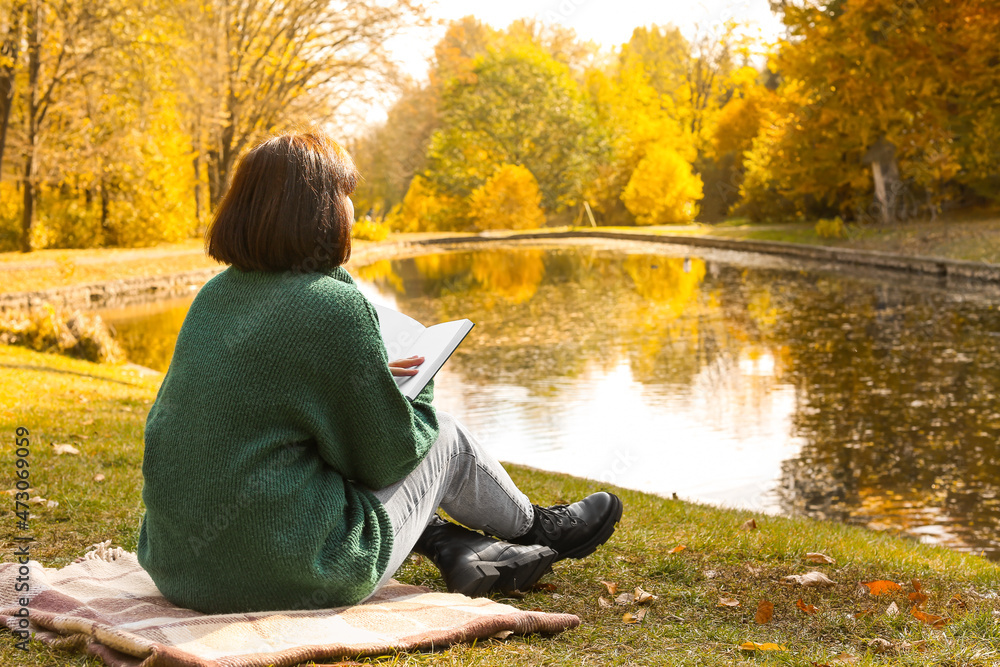 Young woman sitting on plaid and reading book near pond in autumn park