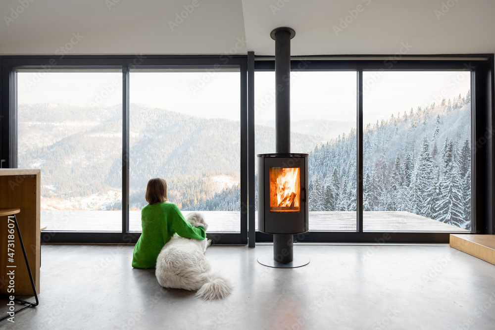 Woman sitting with dog near fireplace and panoramic window at modern living room with stunning view 