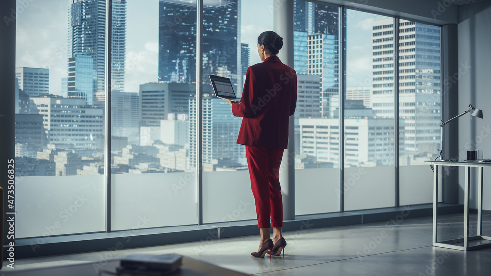 Successful Caucasian Businesswoman Using Laptop While Standing in Office Looking out of Window on Bi