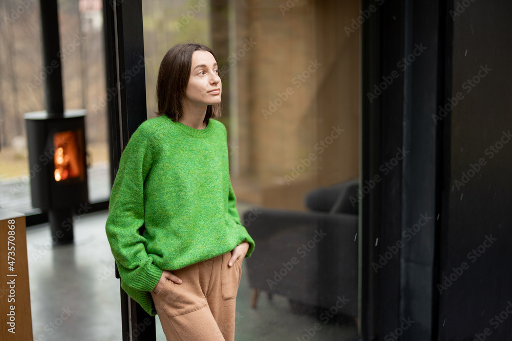 年轻的女人站在现代房子的窗户前，看着大自然。舒适和快乐的概念